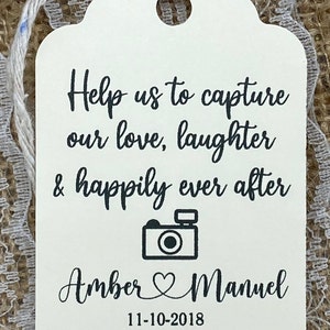 May include: White wedding favor tag with black text that reads "Help us to capture our love, laughter & happily ever after" with a black camera icon and the names "Amber & Manuel" and the date "11-10-2018".
