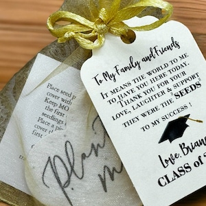 May include: A white card with black text that reads "To My Family and Friends It means the world to me to have you here today. Thank you for your love, laughter & support. They were the "seeds" to my success!"  A black graduation cap with a gold tassel is above the text. Below the text is "Love, Briana Class of 2024". The card is attached to a small, gold mesh bag with a gold ribbon.