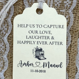 May include: A white wedding favor tag with black text that reads "Help us to capture our love, laughter & happily ever after."  A vintage camera illustration is above the names "Amber & Manuel" and the date "11-10-2018".