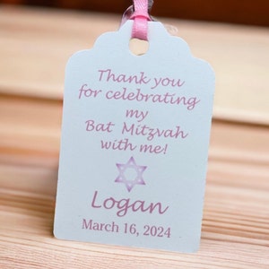 May include: A white thank you tag with pink text and a pink star of David. The text reads "Thank you for celebrating my Bat Mitzvah with me! Logan March 16, 2024".
