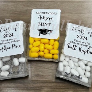 May include: Three clear plastic containers filled with small candies. Each container has a white label with black text that reads "Class of 2024" and a personalized message. The middle container has a yellow graduation cap on the label and the text "Outstanding Achieve Mint".
