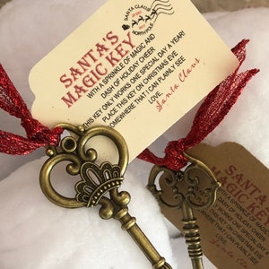 May include: Two antique gold-toned skeleton keys with ornate designs, hanging from a red ribbon. The keys are attached to white gift tags with the text "Santa's Magic Key" and a message about a sprinkle of magic and a dash of holiday cheer.