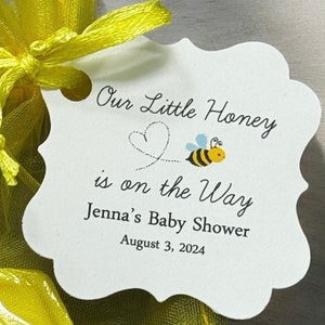 May include: White tag with a bee design and the text "Our Little Honey is on the Way Jenna's Baby Shower August 3, 2024".
