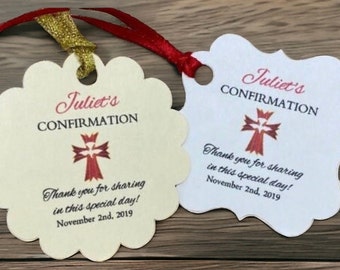 May include: Two white favor tags with scalloped edges. The tags feature a red cross with a dove, the text "Juliet's Confirmation", and the date "November 2nd, 2019". The tags are tied with a gold ribbon.