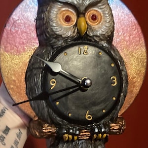 May include: A decorative owl-shaped clock with a black clock face and gold numerals. The owl has gray feathers, orange eyes, and a yellow beak. The clock is mounted on a circular background with pink, gold, and blue hues.