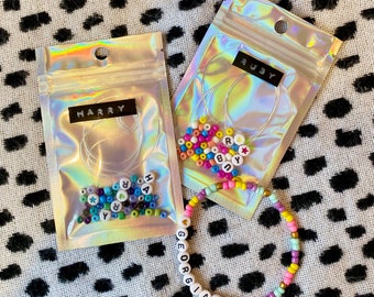 May include: Two clear plastic bags with colorful alphabet beads and string. The bags are labeled "HARRY" and "RUBY". A completed bracelet with the name "GEORGIANA" is shown in the bottom right corner.