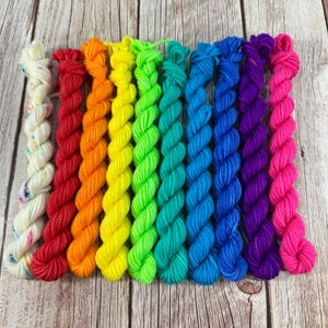 Puede incluir: Una colección de madejas de hilo coloridas dispuestas en un espectro de arcoíris. Las madejas están bien enrolladas y muestran una gama de colores vibrantes, incluyendo rojo, naranja, amarillo, verde, azul, morado y rosa. El hilo se muestra sobre una superficie de madera.