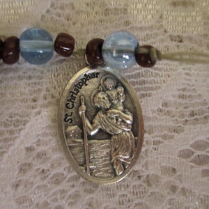 May include: Silver tone St. Christopher medal with a depiction of St. Christopher carrying a child. The medal is attached to a brown and blue beaded necklace.