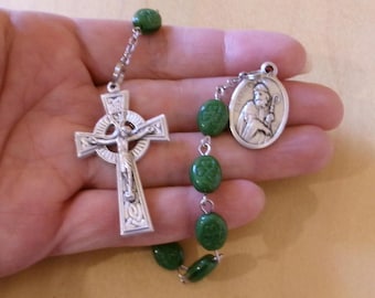 May include: A silver-toned rosary with a Celtic cross and a medallion featuring a religious figure. The rosary beads are oval-shaped and green. The cross depicts Jesus Christ. The rosary is held in a hand.
