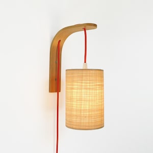 May include: A wall-mounted lamp with a natural wood bracket and a cylindrical lampshade. The lamp features a red cord and a light-colored, textured lampshade. The wooden bracket has a curved design, and the lamp is against a white wall.