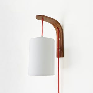 May include: A modern wall-mounted lamp with a white cylindrical shade. The lamp features a curved, wooden bracket and a bright red power cord. The wooden bracket is attached to the wall with screws. The lamp is designed for indoor use.