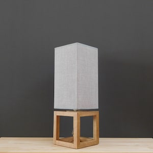 May include: A gray linen lampshade sits atop a wooden cube base. The lamp is on a light wood surface.