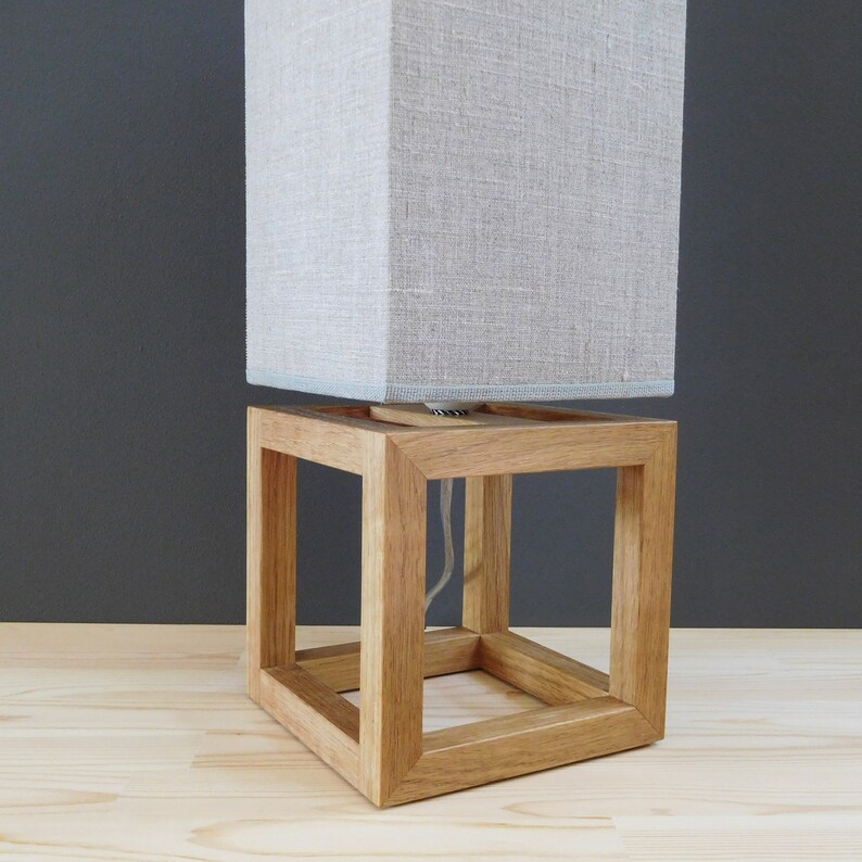 May include: A wooden lamp base with a square, open frame design. The lampshade is made of a light gray fabric with a subtle texture.