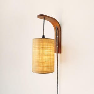 Op de afbeelding: Een houten wandlamp met een gebogen arm en een cilindrische lampenkap. De lampenkap is gemaakt van een lichtgekleurd, getextureerd materiaal en het snoer is grijs en zwart. De lamp is aan een witte muur bevestigd.