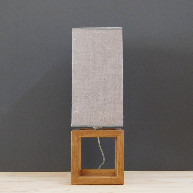 May include: A wooden table lamp with a light beige linen lampshade. The lamp has a square base and a clear cord.