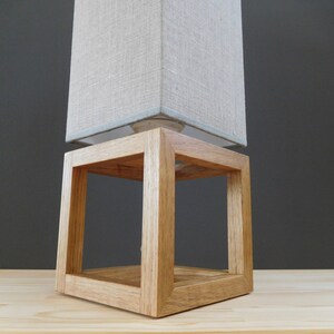 May include: A wooden lamp base with a square, open frame design. The base is made of natural wood and has a light brown finish. A beige linen lampshade sits on top of the base.