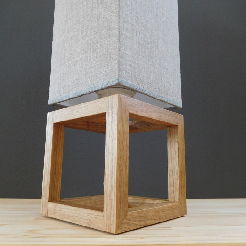 May include: A wooden lamp base with a square, open frame design. The base is made of natural wood and has a light brown finish. A beige linen lampshade sits on top of the base.