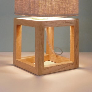 May include: A wooden lamp base with a square shape and a light brown finish. The lampshade is made of a light gray fabric.