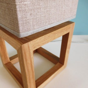 May include: A wooden lamp base with a square shape and a light brown linen lampshade. The base is made of natural wood and has a simple, modern design.