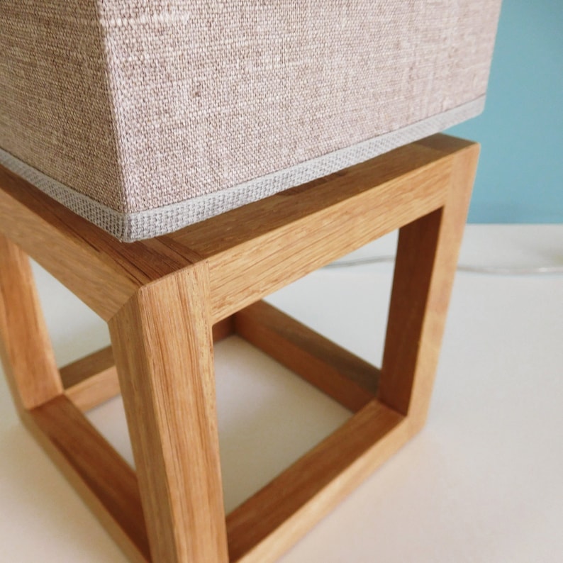 May include: A wooden lamp base with a square shape and a light brown linen lampshade. The base is made of natural wood and has a simple, modern design.