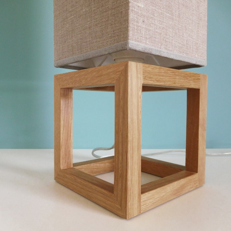 May include: A wooden lamp base with a square, open frame design. The base is made of light brown wood and has a natural finish. The lamp base is designed to hold a lampshade.