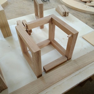 May include: A wooden cube frame is being assembled on a white work surface. The frame is made of light brown wood and has a simple design. The frame is being assembled using dowels and glue.