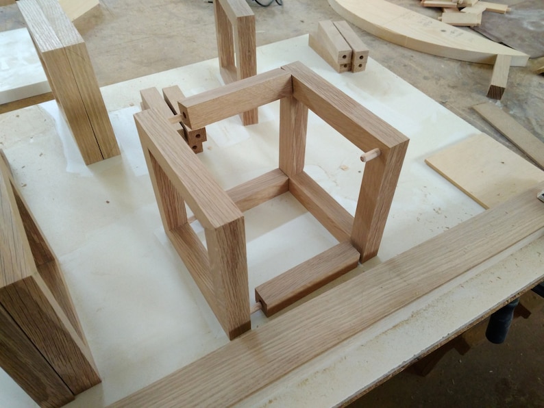 May include: A wooden cube frame is being assembled on a white work surface. The frame is made of light brown wood and has a simple design. The frame is being assembled using dowels and glue.