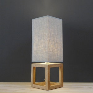 May include: A gray linen lampshade sits atop a wooden cube base. The lampshade is lit, casting a warm glow. The cube base has a square opening on each side.