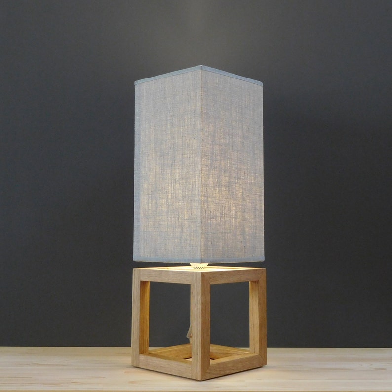 May include: A gray linen lampshade sits atop a wooden cube base. The lampshade is lit, casting a warm glow. The cube base has a square opening on each side.