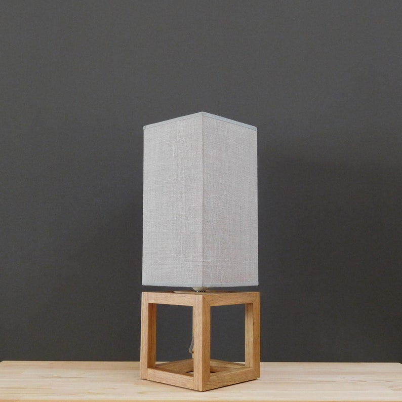 May include: A gray linen lampshade sits atop a wooden cube base. The lamp is on a light wood surface.