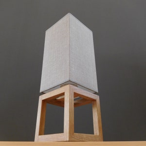 May include: A wooden table lamp with a square linen lampshade. The lampshade is a light beige color and the base is a natural wood color.