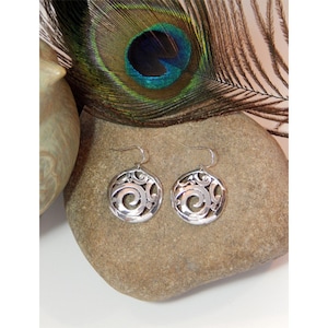 May include: Silver dangle earrings with a circular design featuring a swirl pattern. The earrings are on a gray stone surface.