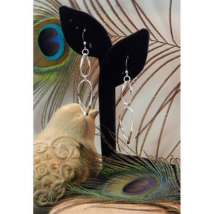 May include: Silver dangle earrings with a twisted, leaf-shaped design. The earrings are displayed on a black velvet stand against a background of peacock feathers.