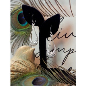 May include: Silver teardrop earrings with a hammered texture, displayed on a black velvet earring stand. The earrings have a simple hook closure. The background includes a decorative bird figurine and peacock feathers.