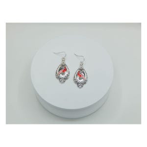 May include: A pair of silver-toned earrings with a decorative, ornate design. Each earring features a red cardinal bird in the center. The earrings have a hook closure for pierced ears. The earrings are displayed on a white surface.