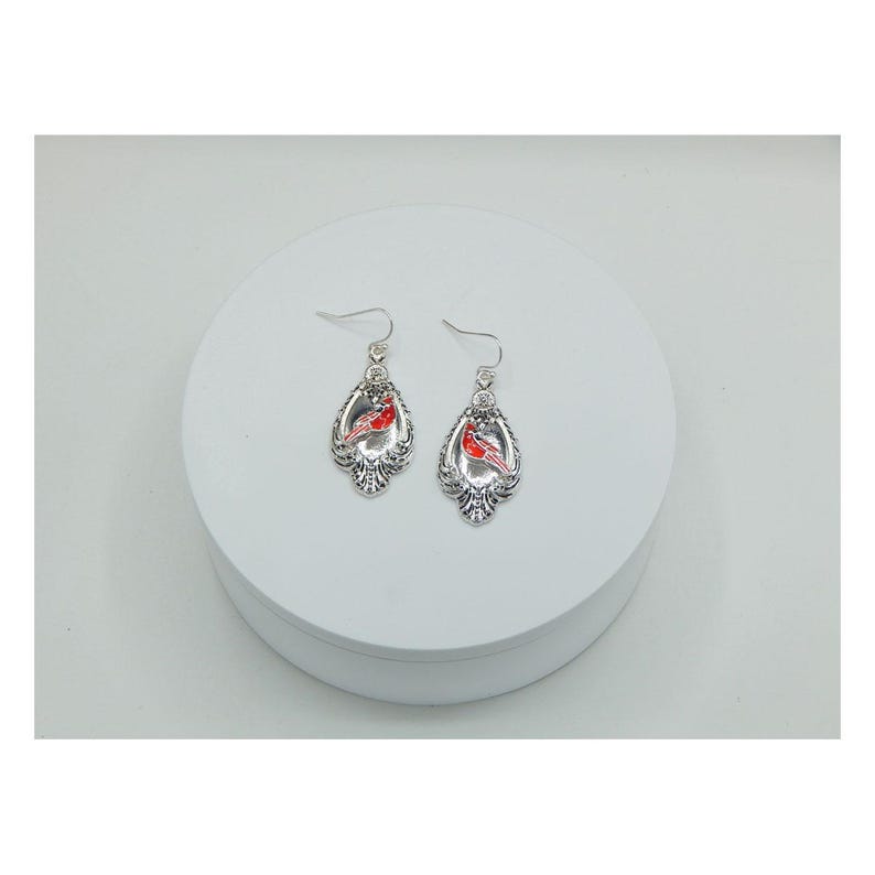 May include: A pair of silver-toned earrings with a decorative, ornate design. Each earring features a red cardinal bird in the center. The earrings have a hook closure for pierced ears. The earrings are displayed on a white surface.
