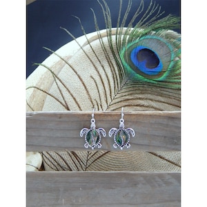 May include: A pair of silver turtle earrings with abalone shell inlays. The earrings are hanging from a wooden surface with a peacock feather in the background.