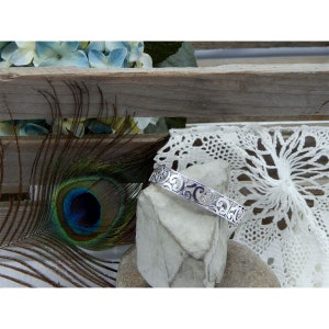 May include: A silver bracelet with a delicate, intricate design. The bracelet is on a gray stone surface with a white lace doily and a peacock feather in the background.