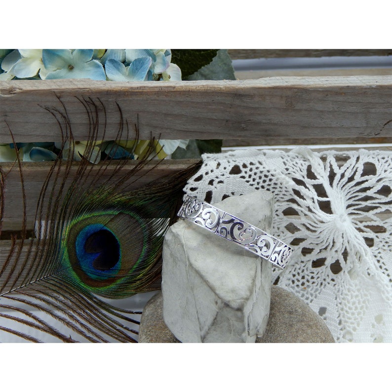 May include: A silver bracelet with a delicate, intricate design. The bracelet is on a gray stone surface with a white lace doily and a peacock feather in the background.