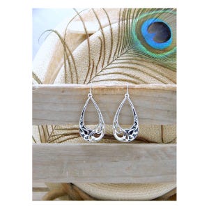 May include: A pair of silver teardrop earrings with an open filigree design. The earrings are displayed on a wooden surface with a peacock feather in the background. The earrings have a delicate, ornate pattern.