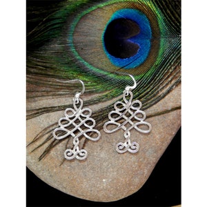 May include: A pair of silver-colored earrings with an intricate, swirling design. The earrings are displayed on a light brown stone, with a peacock feather in the background. The earrings have a hook closure.