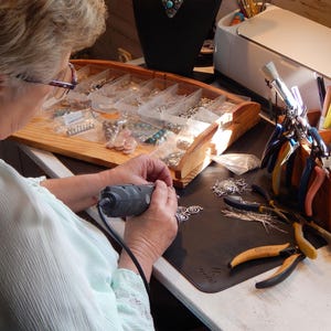 May include: A person working on jewelry, using a rotary tool. The workspace includes a wooden organizer with jewelry components, tools, and a variety of pliers. The scene suggests a jewelry-making studio.