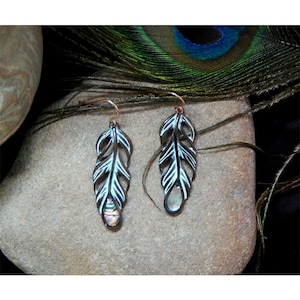 May include: A pair of feather-shaped earrings. The earrings are dark metal with white accents and a pearlescent stone at the bottom. The earrings hang from copper-colored hooks. The background includes a peacock feather.