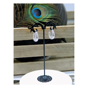 May include: A pair of silver hoop earrings with intricate openwork designs are displayed on a black earring stand. The stand is on a white surface, with a peacock feather and wooden background.