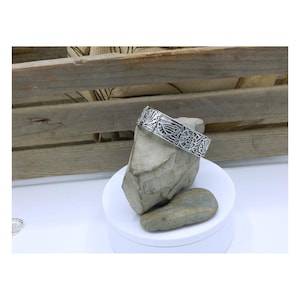 May include: A silver-toned bracelet with an intricate, openwork design featuring floral and geometric patterns. The bracelet is displayed on a gray rock, set against a wooden crate background. The jewelry is a statement piece.