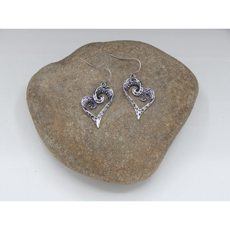 Puede incluir: Un par de pendientes de plata en forma de coraz&oacute;n con un dise&ntilde;o de espiral. Los pendientes cuelgan de una roca.