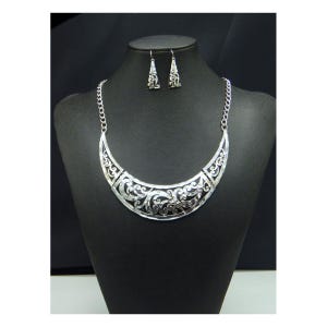 May include: A silver-toned necklace and earring set displayed on a black mannequin. The necklace features a crescent-shaped pendant with intricate scrollwork. The matching earrings have a similar design. The jewelry is likely made of metal.