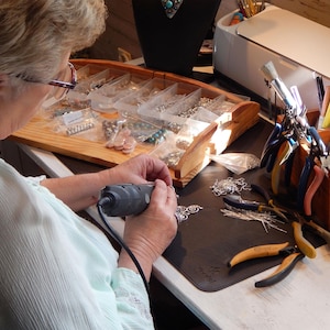 May include: A person working on jewelry making with a variety of tools and beads. The person is using a small electric drill to work on a piece of jewelry. There are many beads and tools on the table, including pliers, wire cutters, and a small container of beads.