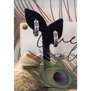 May include: A pair of silver hoop earrings with an intricate floral design. The earrings are displayed on a black velvet stand.
