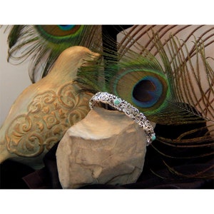 May include: A silver-toned bracelet with an ornate, swirling design and small turquoise-colored stones. The bracelet is displayed on a stone, with a decorative bird statue and peacock feathers in the background.